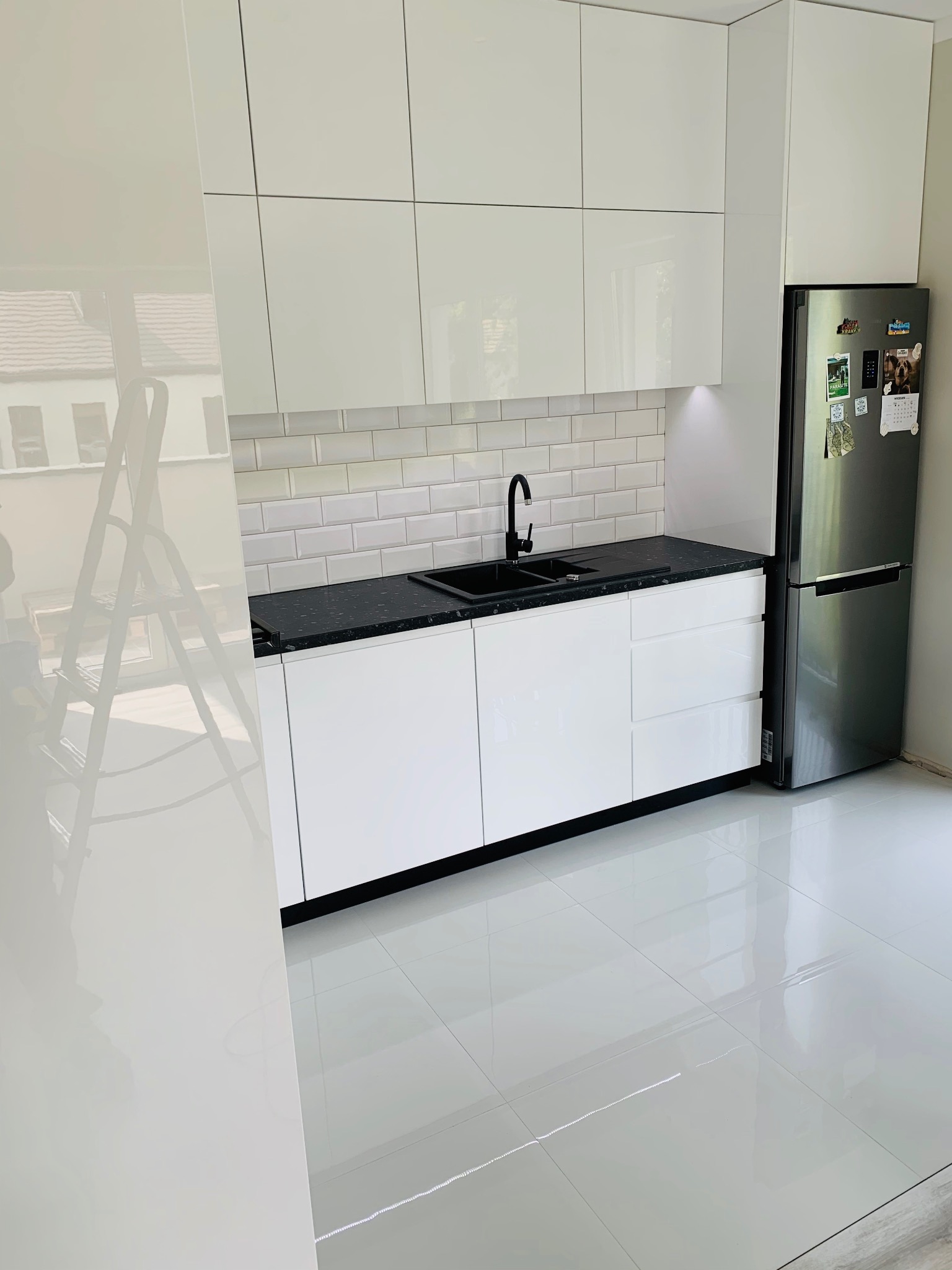 Nowoczesna, jasna kuchnia z białymi lakierowanymi frontami szafek, czarnym blatem i baterią, oraz białymi płytkami na ścianie. W tle widoczna drabina malarska odbijająca się w szafce.