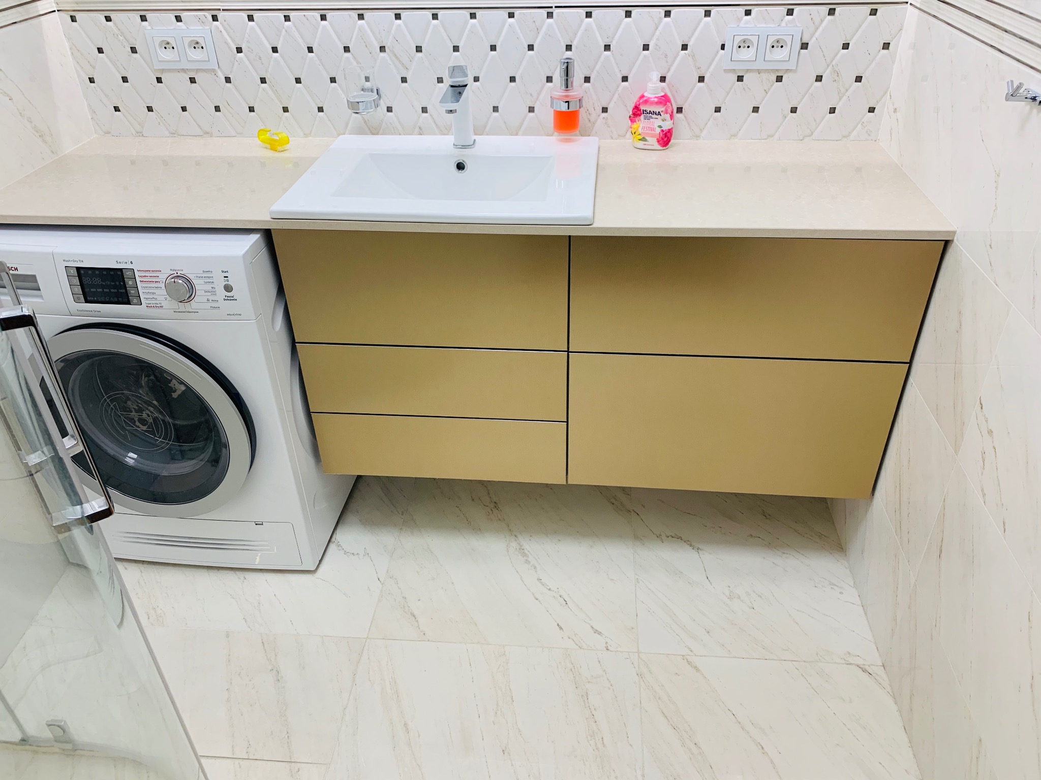 Nowoczesna łazienka z beżową szafką pod umywalką, białą umywalką, pralką Bosch Serie 6 i marmurowymi płytkami na ścianach i podłodze.