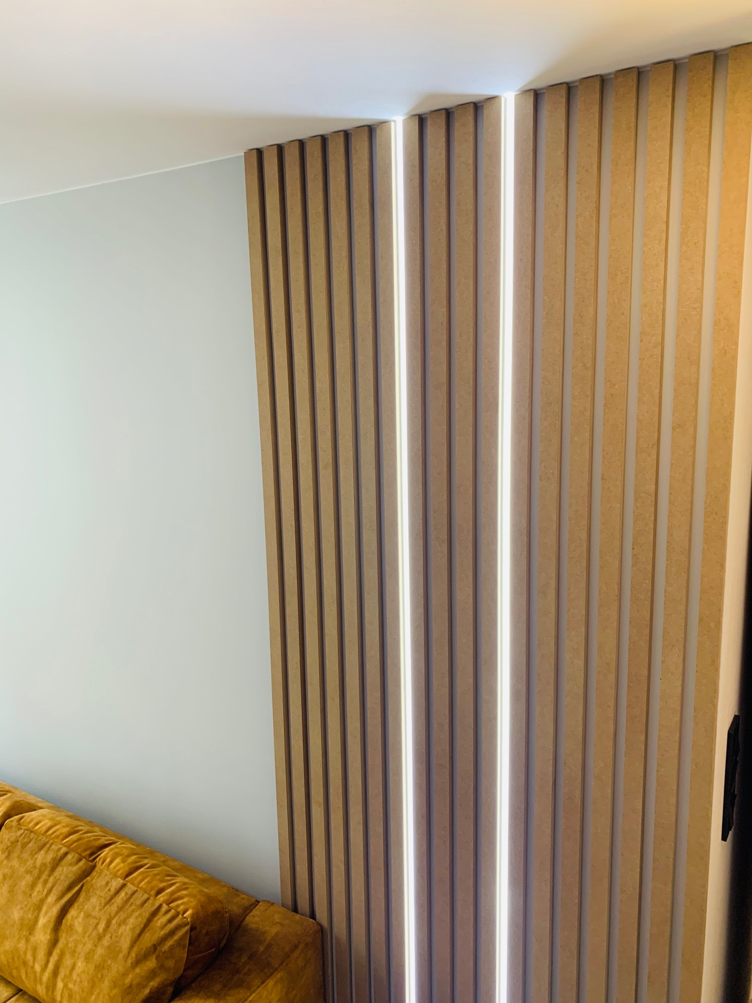 Panel ścienny z pionowych listewek z podświetleniem LED, widoczny fragment sofy w kolorze musztardowym, jasna ściana i sufit.