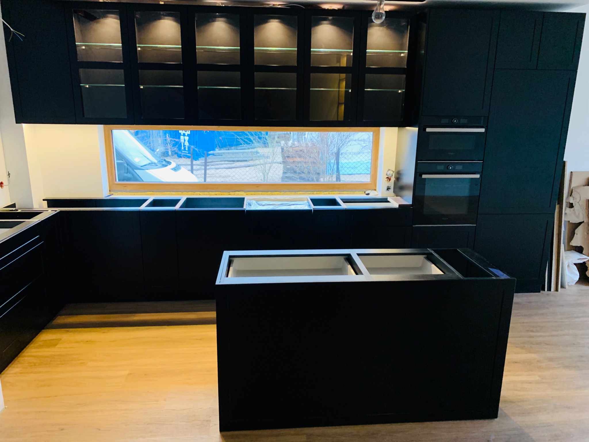 Częściowo zamontowana, nowoczesna kuchnia z czarnymi szafkami, przeszklonymi górnymi szafkami z oświetleniem wewnętrznym i wyspą kuchenną bez blatu, widoczna instalacja okienna i sprzęt AGD.