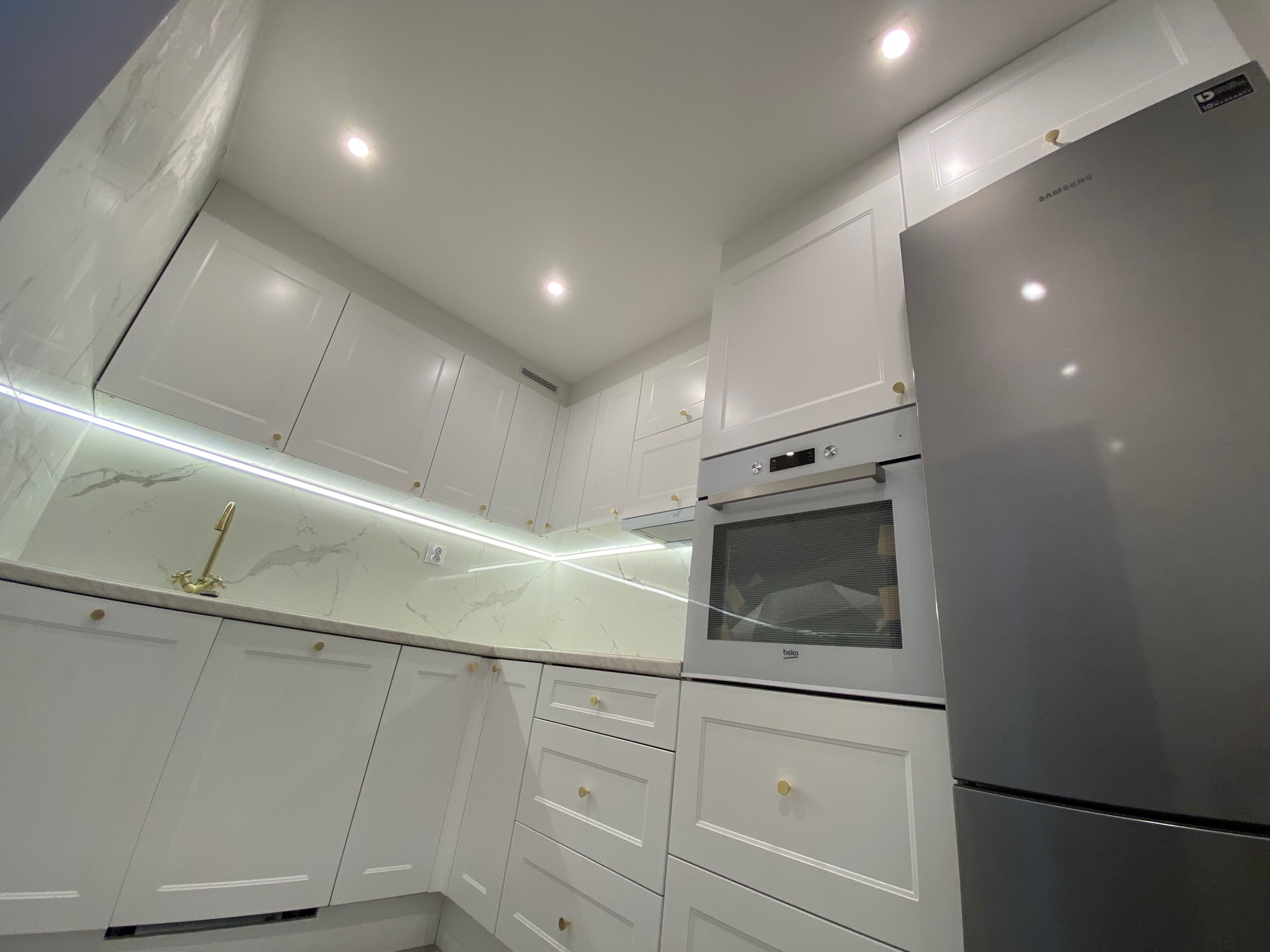 Nowoczesna, jasna kuchnia z białymi szafkami z frezowanymi frontami i złotymi gałkami, marmurowym blatem i podświetleniem LED, uzupełniona srebrną lodówką Samsung i piekarnikiem Beko.