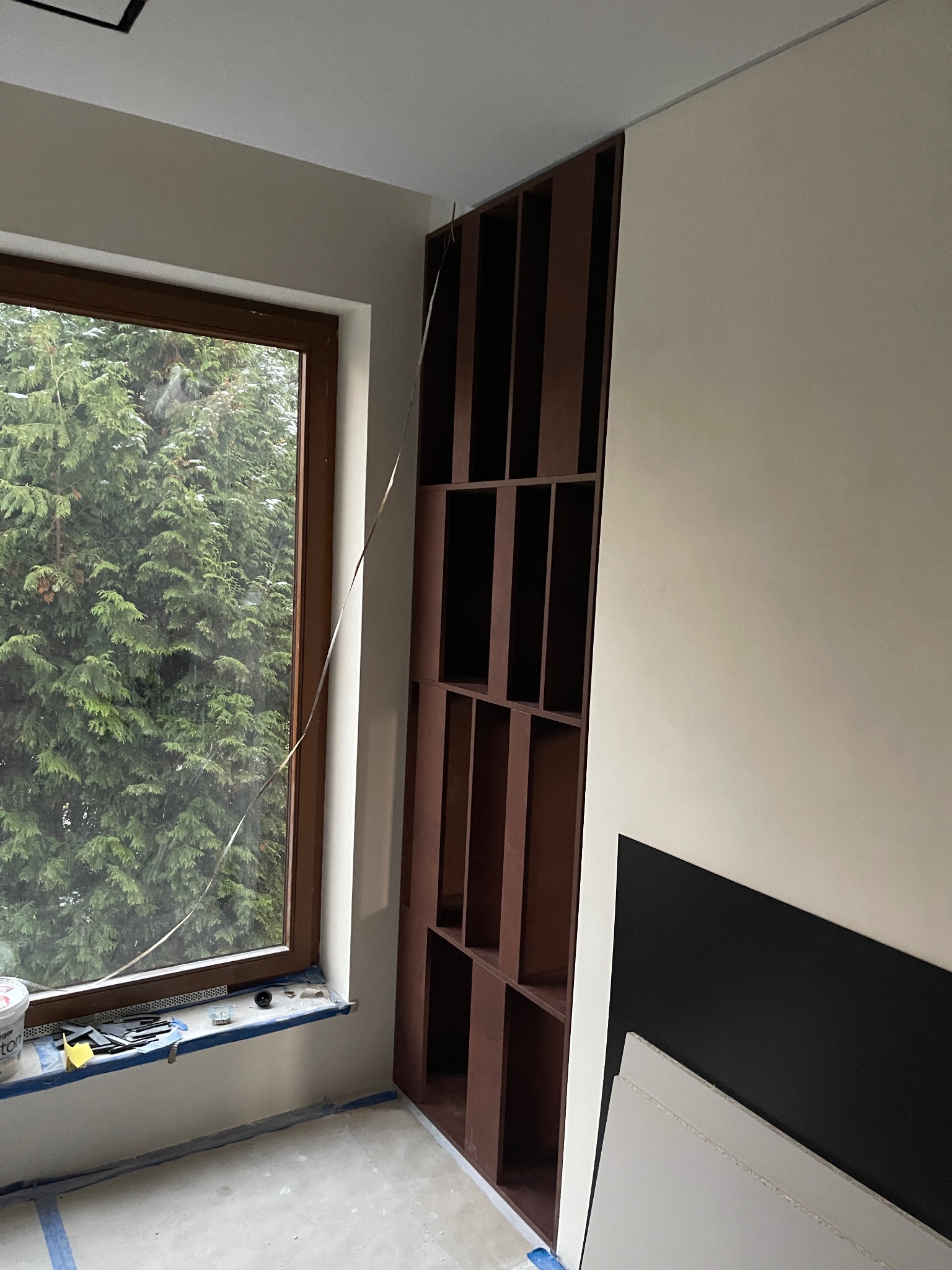 Wnętrze w trakcie wykończenia: brązowy regał wnękowy z otwartymi półkami, okno z widokiem na zielone drzewo, narzędzia malarskie na parapecie, zabezpieczona podłoga.