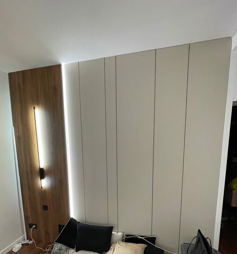 Nowoczesna zabudowa ściany z drewnianym panelem i pionowym oświetleniem LED, widoczne poduszki na podłodze.