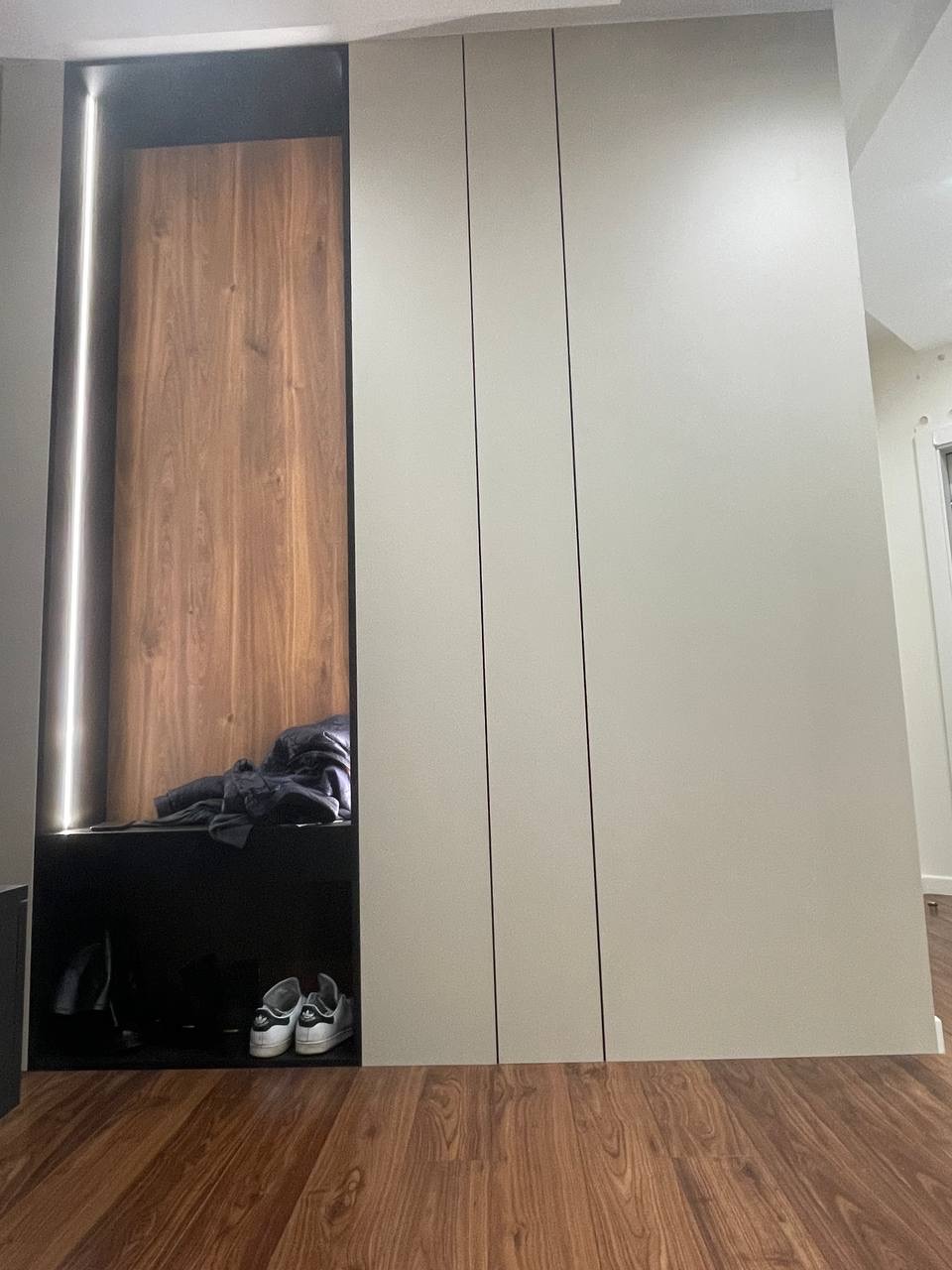 Wnęka z drewnianym panelem i podświetleniem LED sąsiadująca z trzydrzwiową szafą w zabudowie, na podłodze brązowe panele.