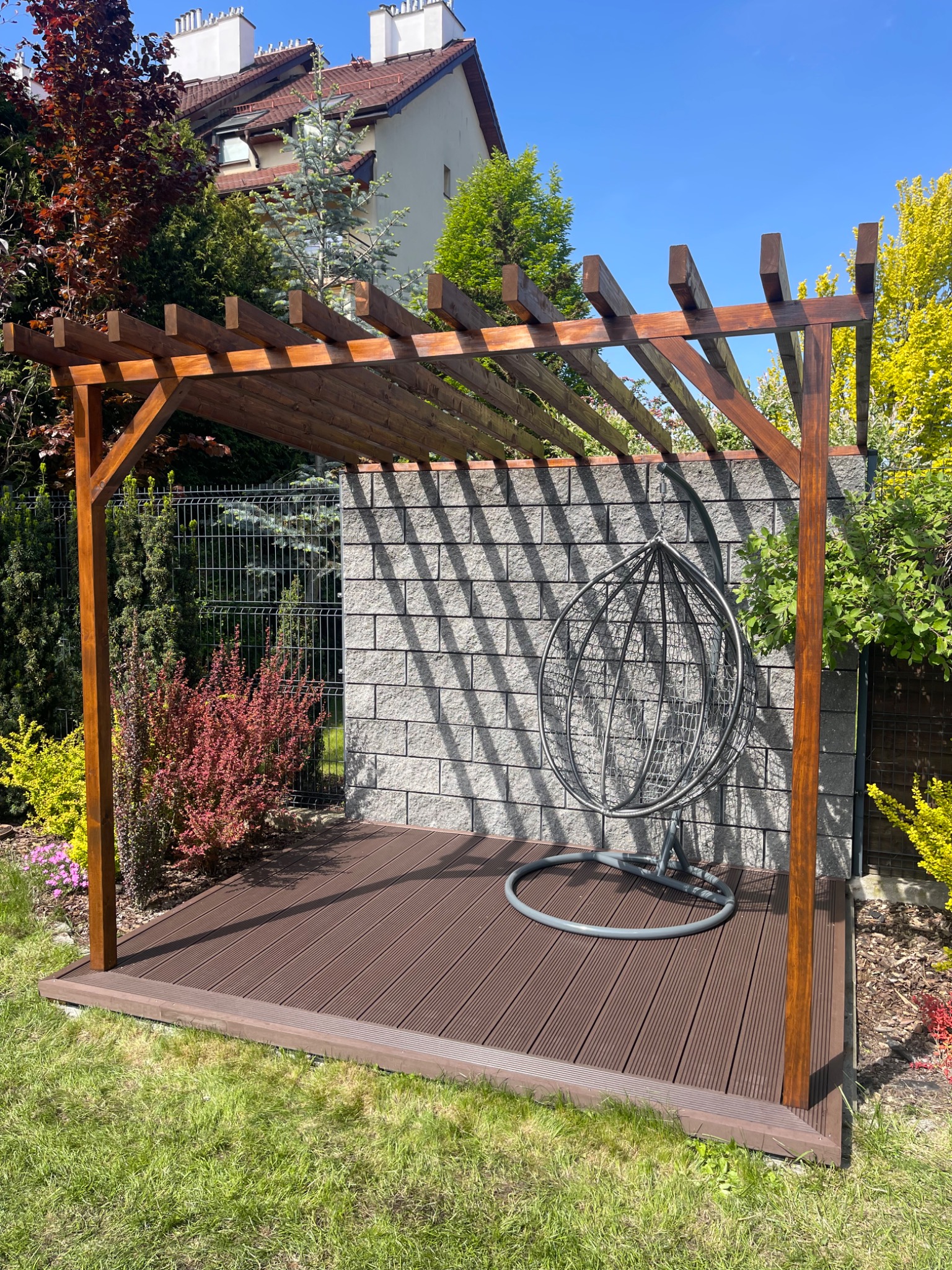 Drewniana pergola z tarasem kompozytowym i wiszącym fotelem. Widok na ogród, szarą ścianę z bloków i metalowe ogrodzenie. Słoneczny dzień.