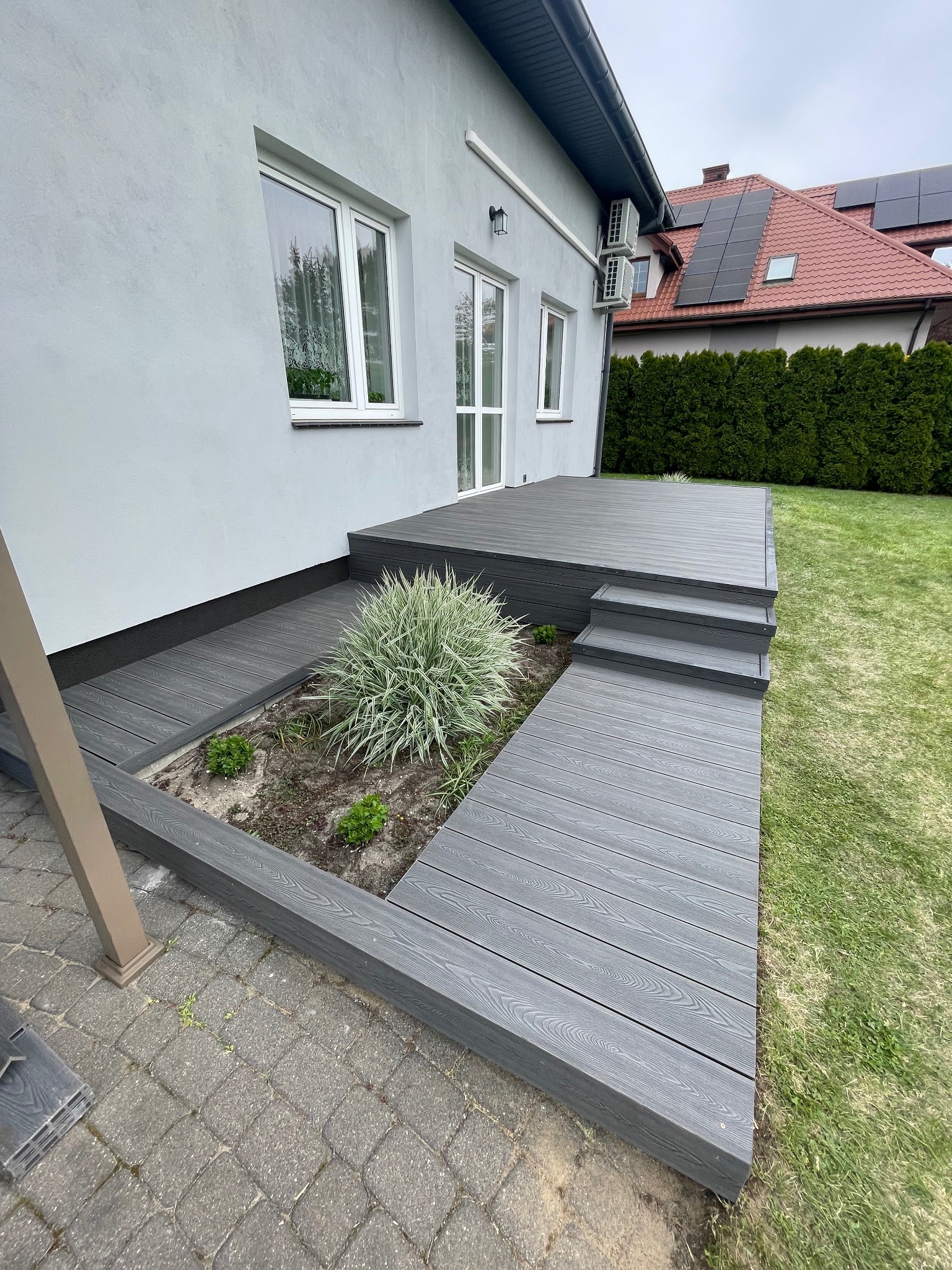 Szary taras kompozytowy z trzema schodami, rabatą z roślinami i widokiem na dom z szarą elewacją i panele słoneczne na dachu w tle.