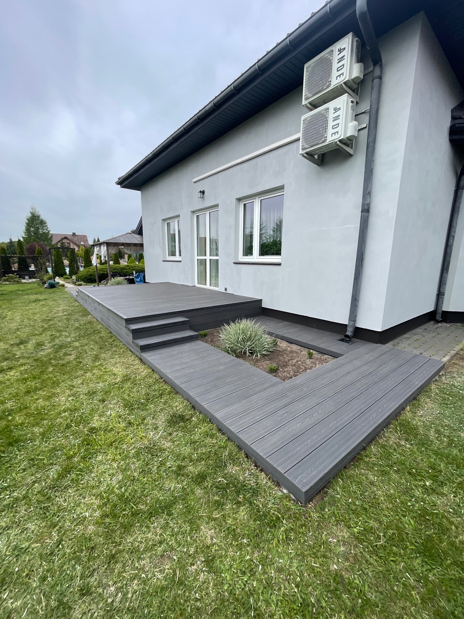 Szary taras kompozytowy z trzema schodkami, otaczający rabatę z rośliną, przylegający do jasnego budynku z dwoma klimatyzatorami na ścianie.