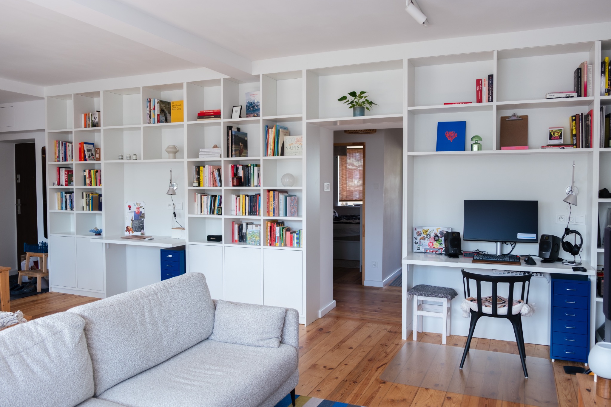 Biały regał na książki z wbudowanymi biurkami w jasnym pokoju dziennym. Widoczny szary narożnik, drewniana podłoga i dekoracje na półkach.