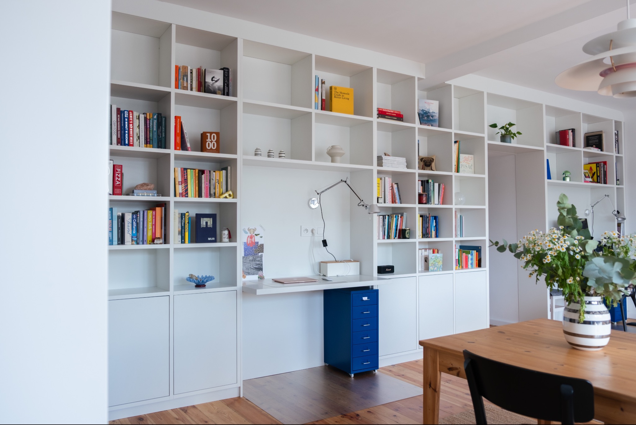 Białe regały na książki z wbudowanym biurkiem i niebieską szafką z szufladami. Na stole wazon z kwiatami. Jasne wnętrze z drewnianą podłogą.