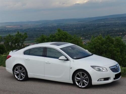 Biała limuzyna Opel Insignia z czarnym dachem panoramicznym, zaparkowana na skraju drogi z widokiem na pagórkowaty krajobraz w tle, w pochmurny dzień.