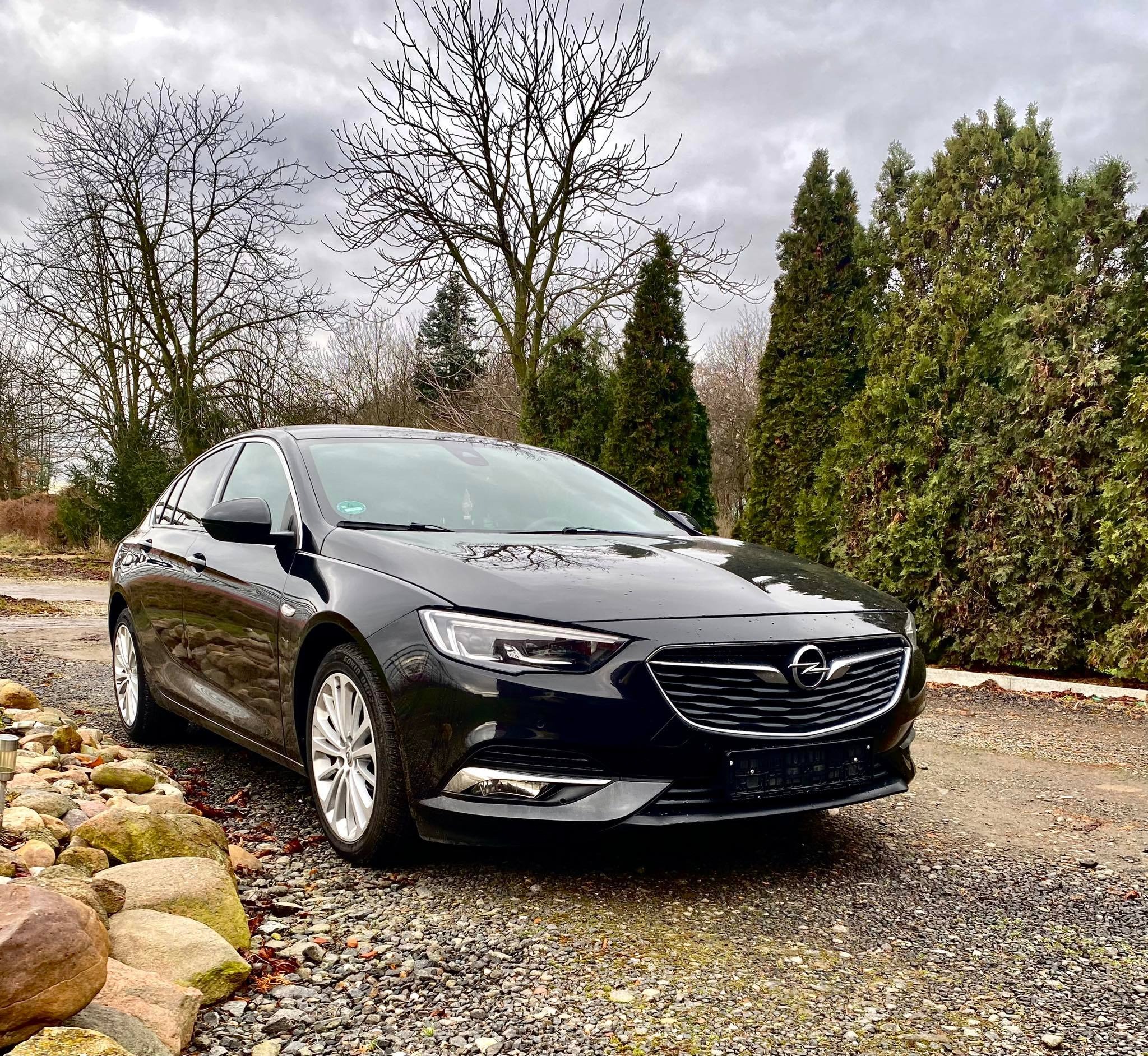 Czarny samochód osobowy marki Opel Insignia zaparkowany na żwirowym podłożu, z widocznymi drzewami iglastymi i bezlistnymi w tle, pod pochmurnym niebem.