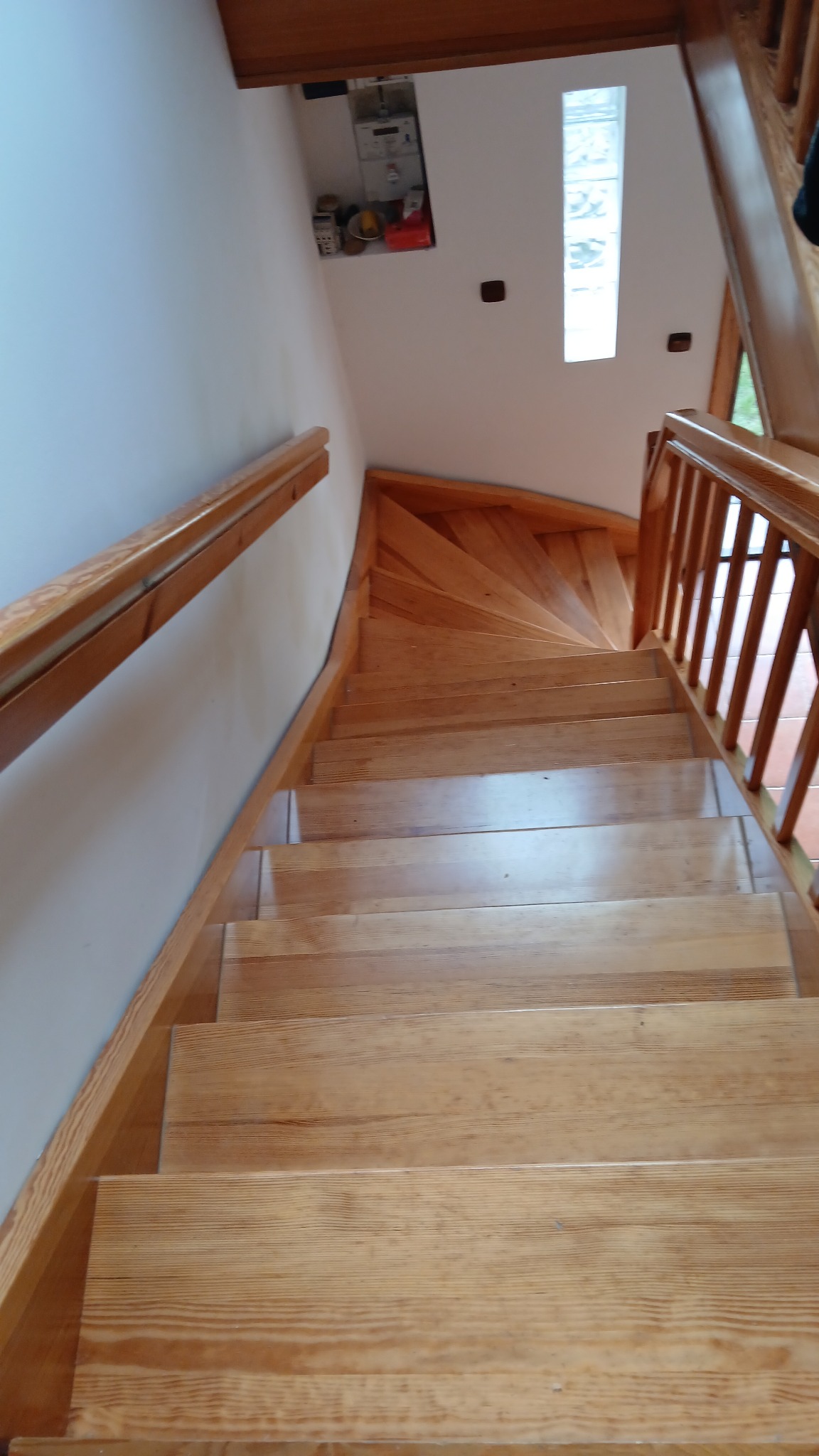 Jasne, drewniane schody z poręczą, widziane z góry. Widoczne naturalne usłojenie drewna i skosy stopni. Biała ściana z elementami wyposażenia w tle.