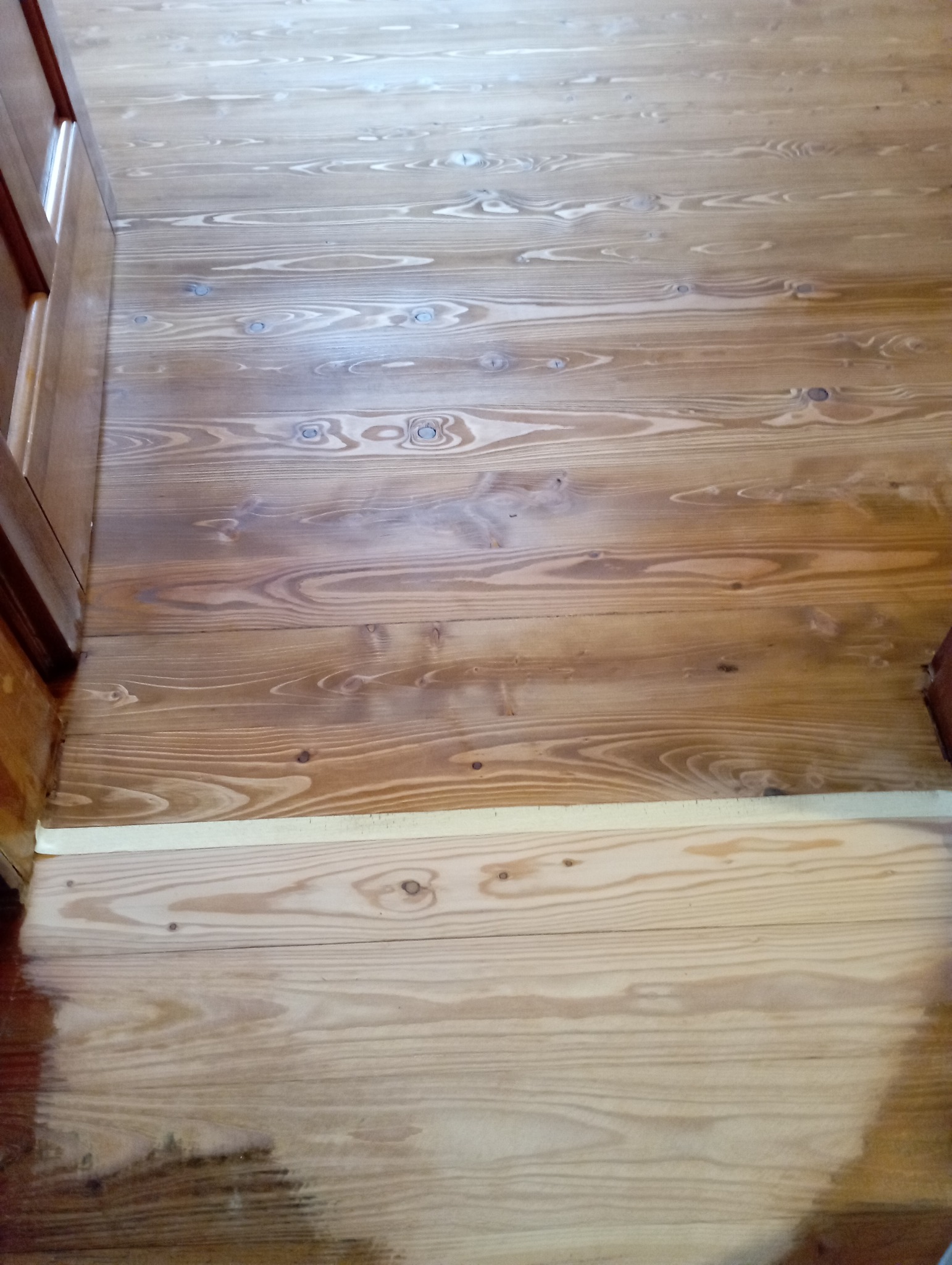 Fragment drewnianej podłogi po częściowym lakierowaniu, widoczne różnice w odcieniach drewna. Taśma malarska zabezpiecza dolną część podłogi.