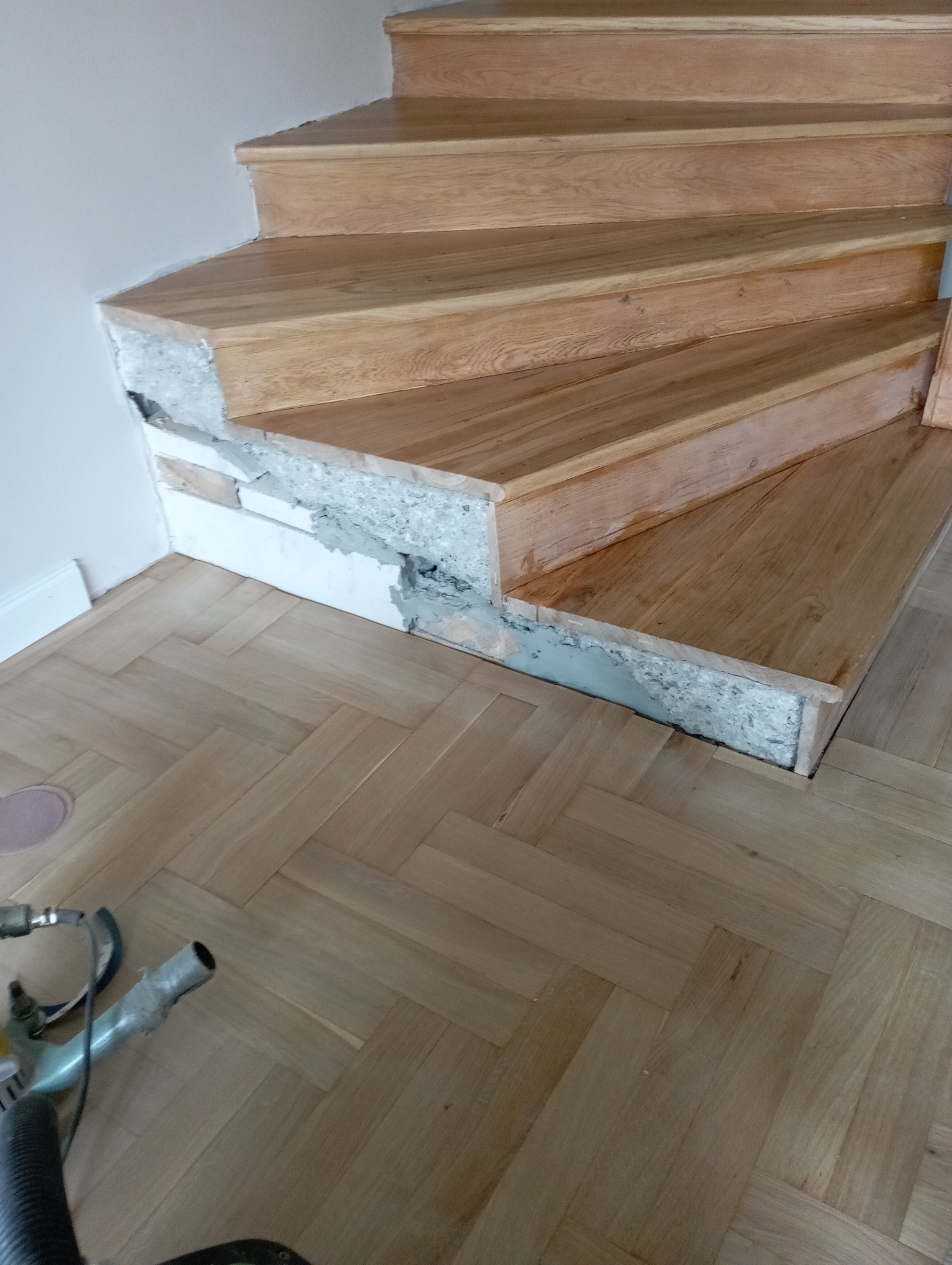 Cyklinowanie schodów z drewna dębowego, widoczne uszkodzenia i naprawy betonu. Parkiet dębowy ułożony w jodełkę. Szlifierka do parkietu w lewym dolnym rogu.