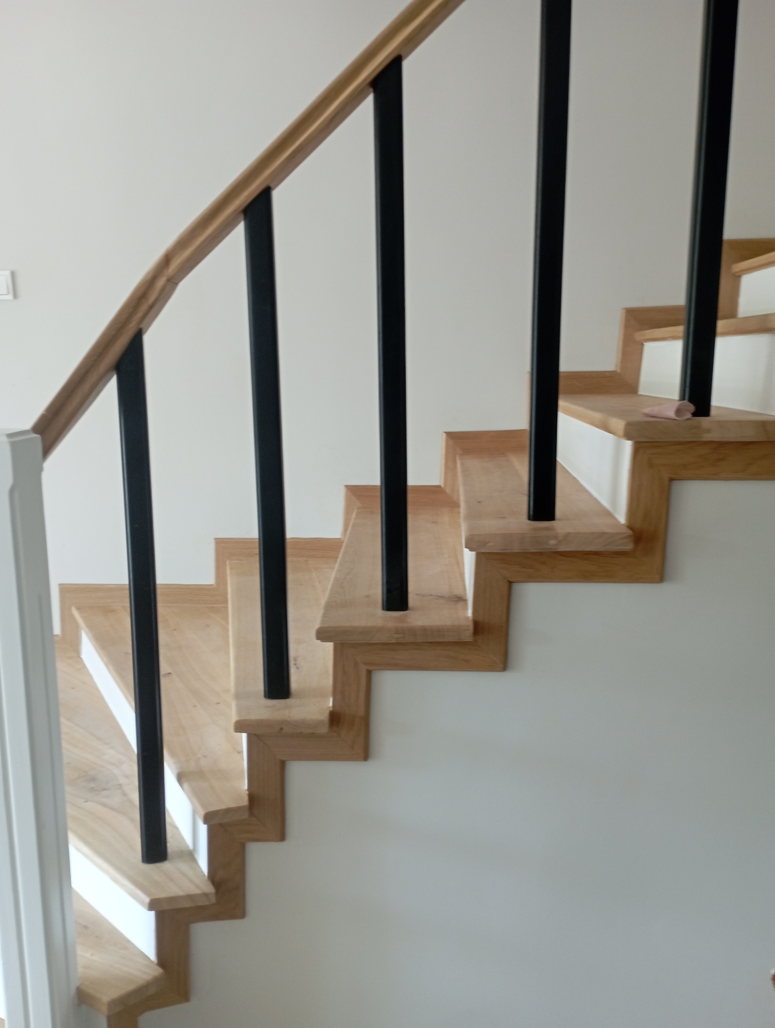 Jasne drewniane schody z czarnymi balustradami i białymi podstopnicami, widok z dołu, minimalistyczny design, naturalne światło.