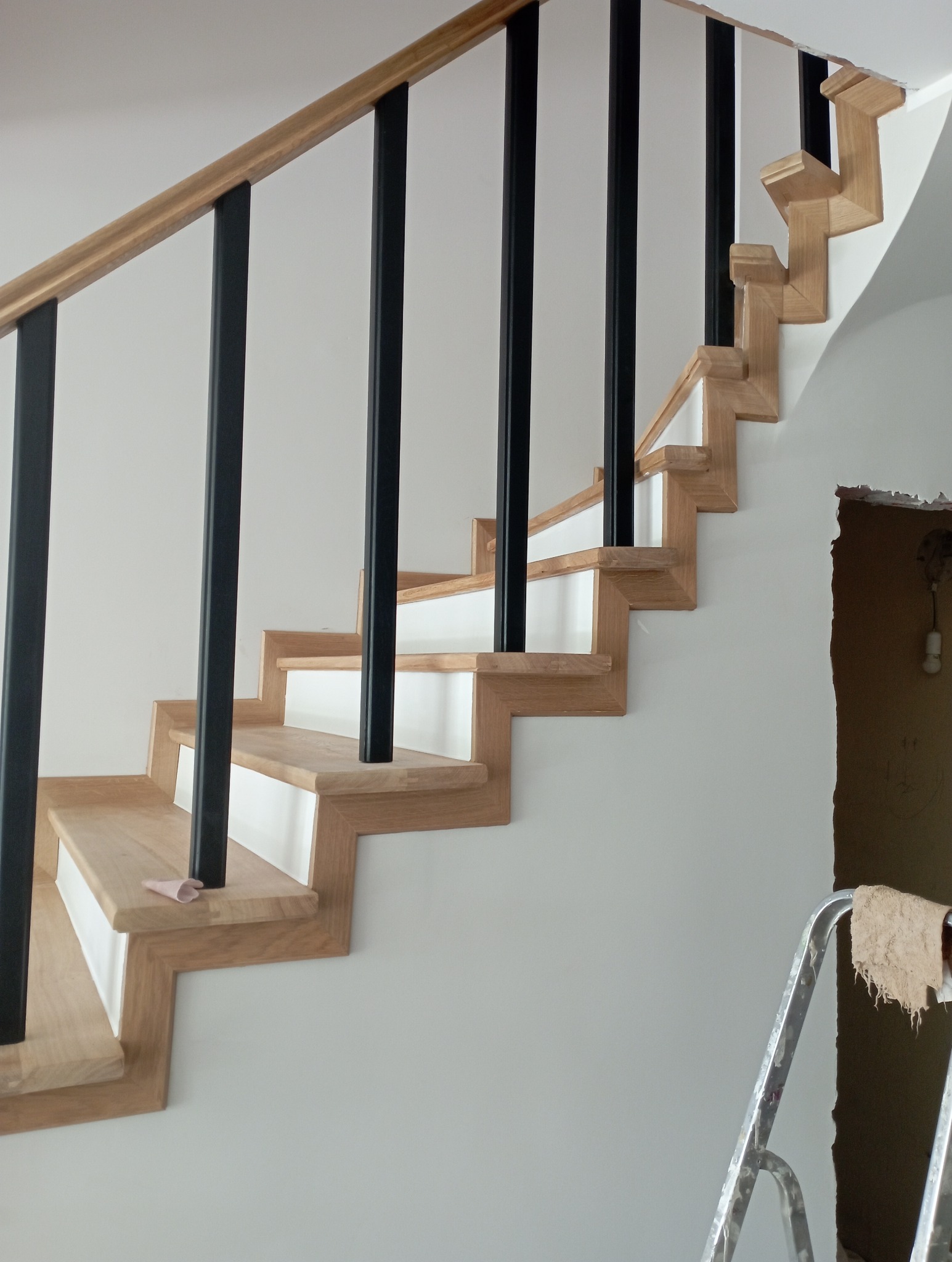 Drewniane schody z czarną balustradą na białym tle. Widoczny fragment drabiny malarskiej i otwór w ścianie. Jasne drewno i nowoczesny design.