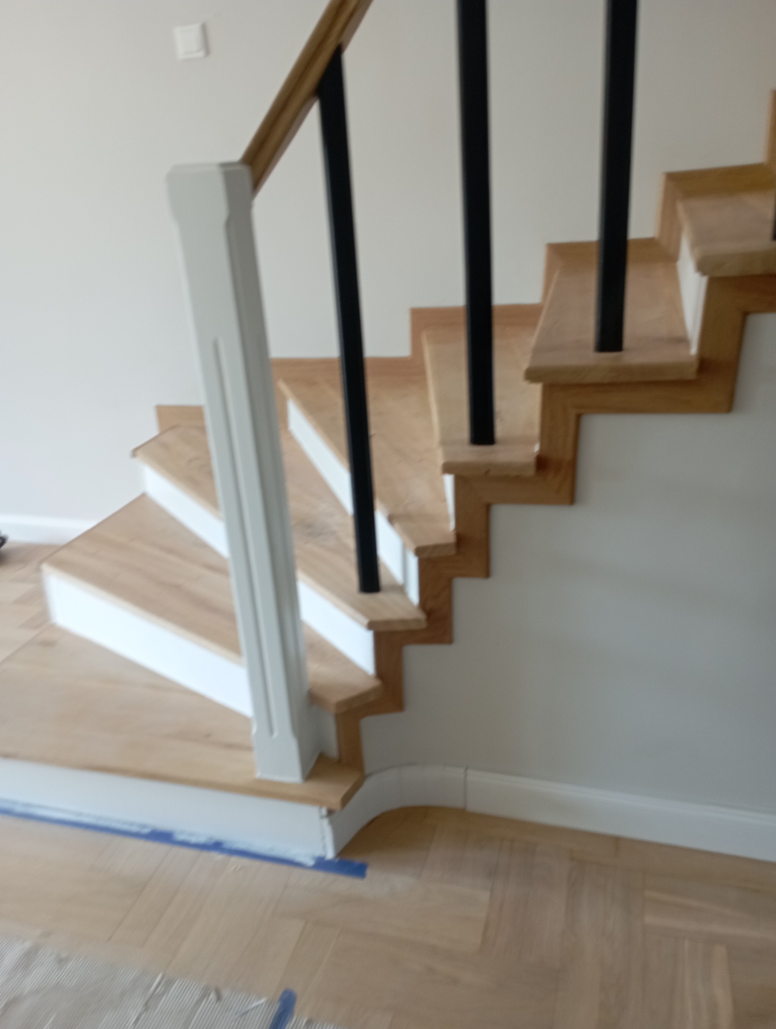 Jasne, drewniane schody z czarną balustradą i białymi listwami przypodłogowymi, wykończone na wysoki połysk. Parkiet ułożony w jodełkę. Widok z dołu.