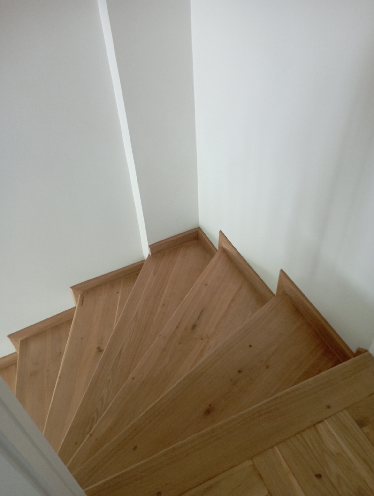 Jasne, drewniane schody z widocznymi słojami drewna, prowadzące w dół, z białymi ścianami po bokach. Ujęcie z góry, podkreślające naturalny wygląd drewna.