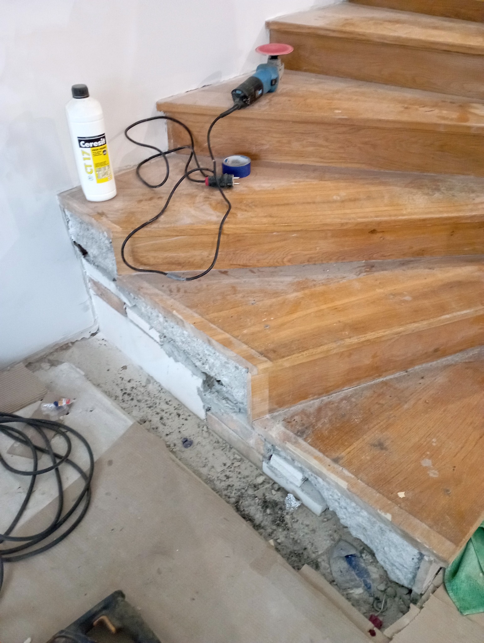 Cyklinowanie drewnianych schodów w trakcie renowacji. Widoczna szlifierka, butelka z preparatem i uszkodzone fragmenty stopni przed naprawą.