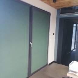 GRZEGORZ PYĆ ATOS-SERWIS ROLET - Dwa okna z wewnętrznymi roletami w kolorze stonowanej zieleni, zamontowane w nowoczesnym wnętrzu z drewnianym sufitem i szarymi ścianami.