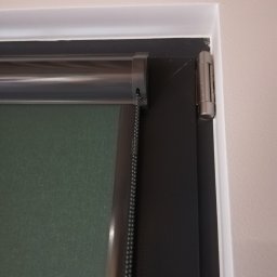 GRZEGORZ PYĆ ATOS-SERWIS ROLET - Fragment okna z zamontowaną roletą wewnętrzną w kolorze ciemnej zieleni, widoczny mechanizm łańcuszkowy i srebrna listwa mocująca.