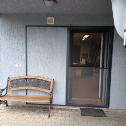 GRZEGORZ PYĆ ATOS-SERWIS ROLET - Szara elewacja domu z zamontowaną moskitierą w drzwiach przesuwnych i drewnianą ławką z motywem winogron.