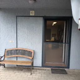 Szara elewacja domu z zamontowaną moskitierą w drzwiach przesuwnych i drewnianą ławką z motywem winogron.