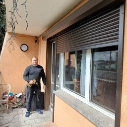 GRZEGORZ PYĆ ATOS-SERWIS ROLET - Pracownik z wiertarką montuje brązową roletę zewnętrzną na balkonie z dekoracyjnymi lampkami.