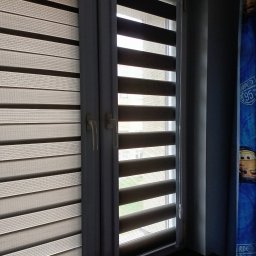 GRZEGORZ PYĆ ATOS-SERWIS ROLET - Dwa okna z zamontowanymi roletami typu dzień i noc w kolorach szarości, jedna z łańcuszkiem sterującym, widok z wnętrza pokoju dziecięcego.