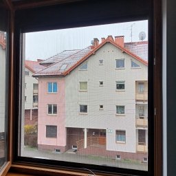 GRZEGORZ PYĆ ATOS-SERWIS ROLET - Zewnętrzna roleta okienna w kolorze grafitowym, częściowo opuszczona, widok z wnętrza na budynek mieszkalny w pochmurny dzień.