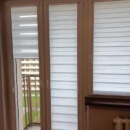 Trzy okna z białymi roletami dzień i noc, widok na balkon i budynek naprzeciwko.