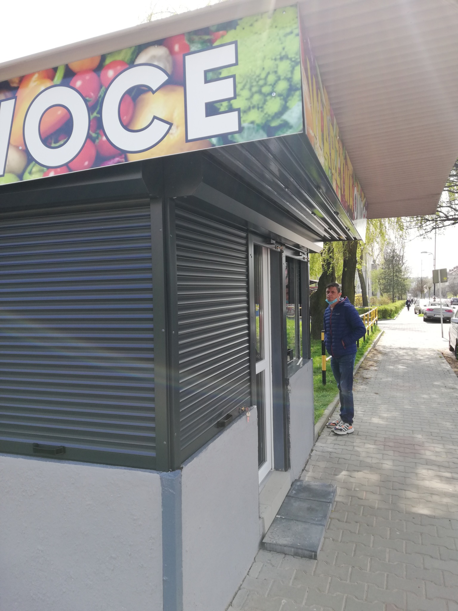 Zamknięty kiosk z owocami i warzywami zabezpieczony szarą roletą zewnętrzną, mężczyzna w maseczce stoi obok.