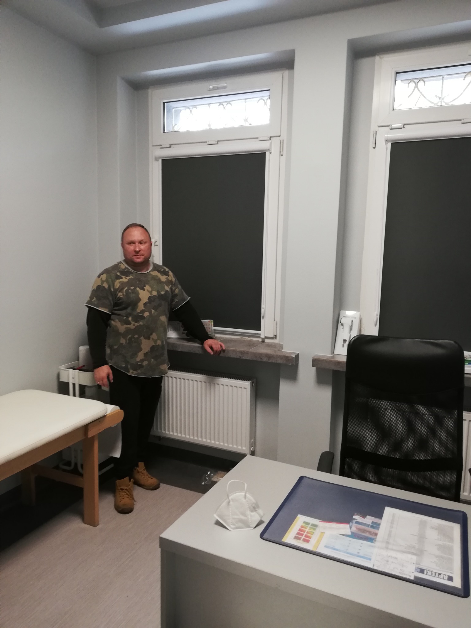Mężczyzna w kamuflażowej koszulce stoi w gabinecie lekarskim przy oknie z zasuniętą roletą w kolorze antracytowym.