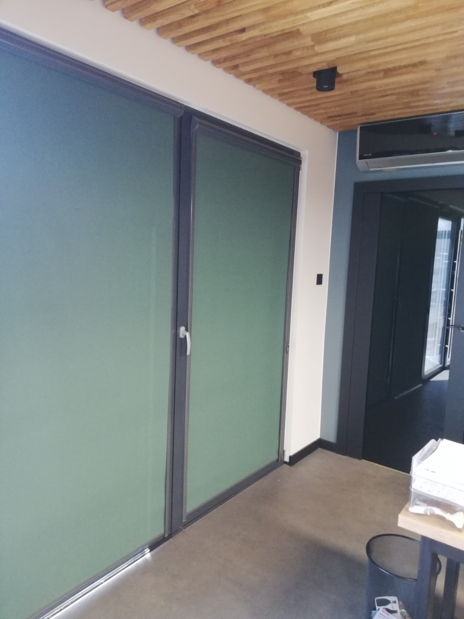 Dwa okna z wewnętrznymi roletami w kolorze stonowanej zieleni, zamontowane w nowoczesnym wnętrzu z drewnianym sufitem i szarymi ścianami.