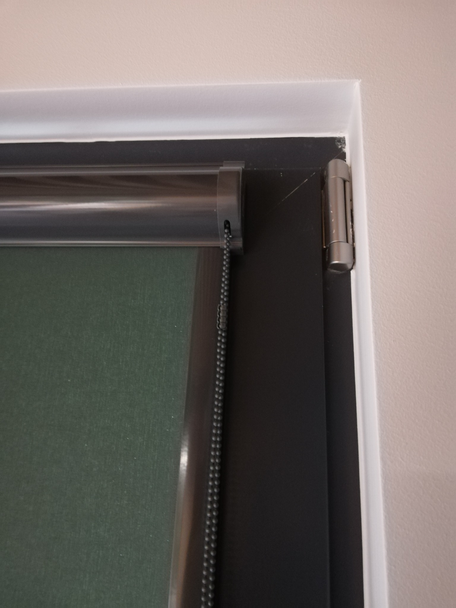 Fragment okna z zamontowaną roletą wewnętrzną w kolorze ciemnej zieleni, widoczny mechanizm łańcuszkowy i srebrna listwa mocująca.