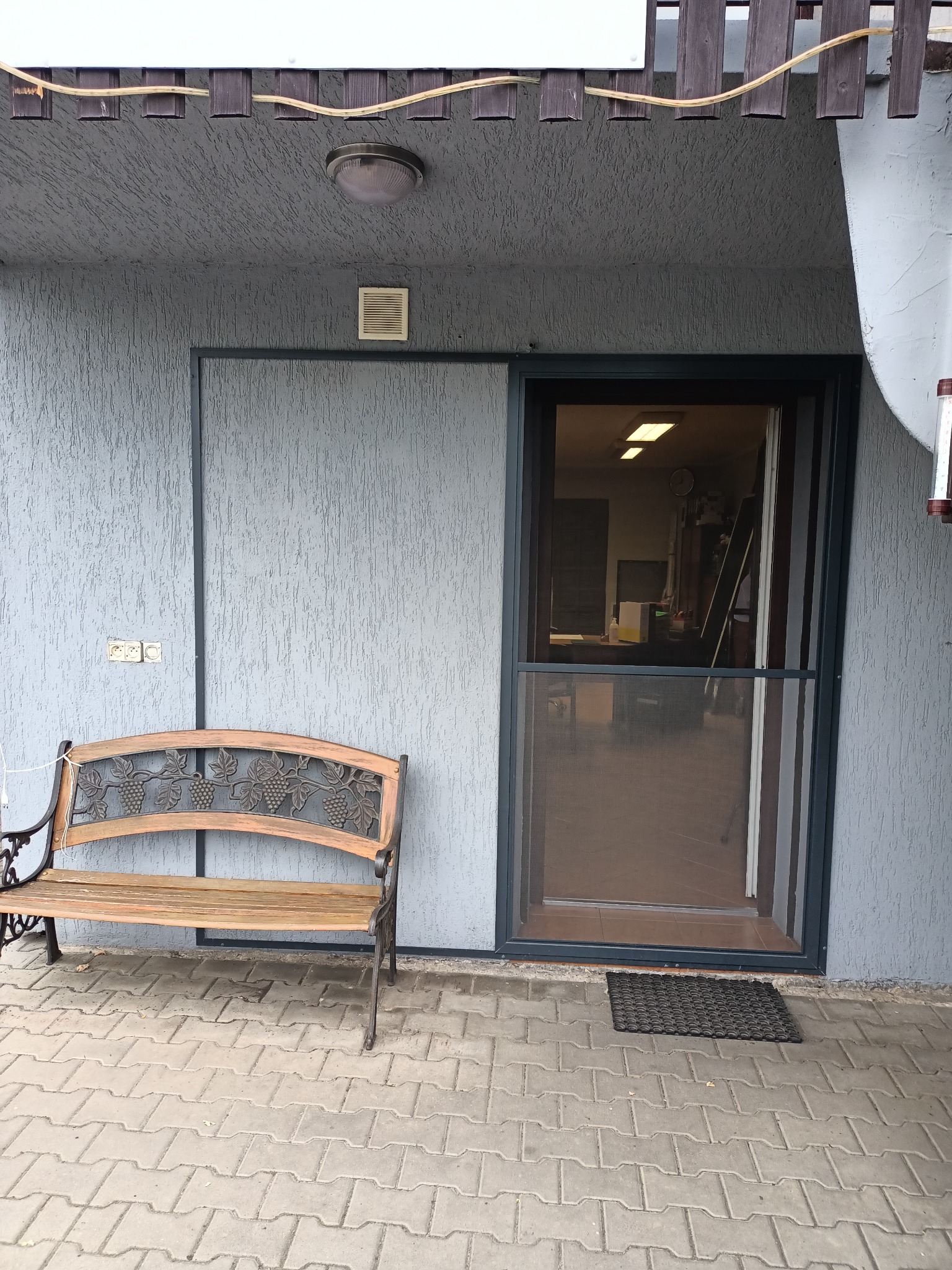 Szara elewacja domu z zamontowaną moskitierą w drzwiach przesuwnych i drewnianą ławką z motywem winogron.