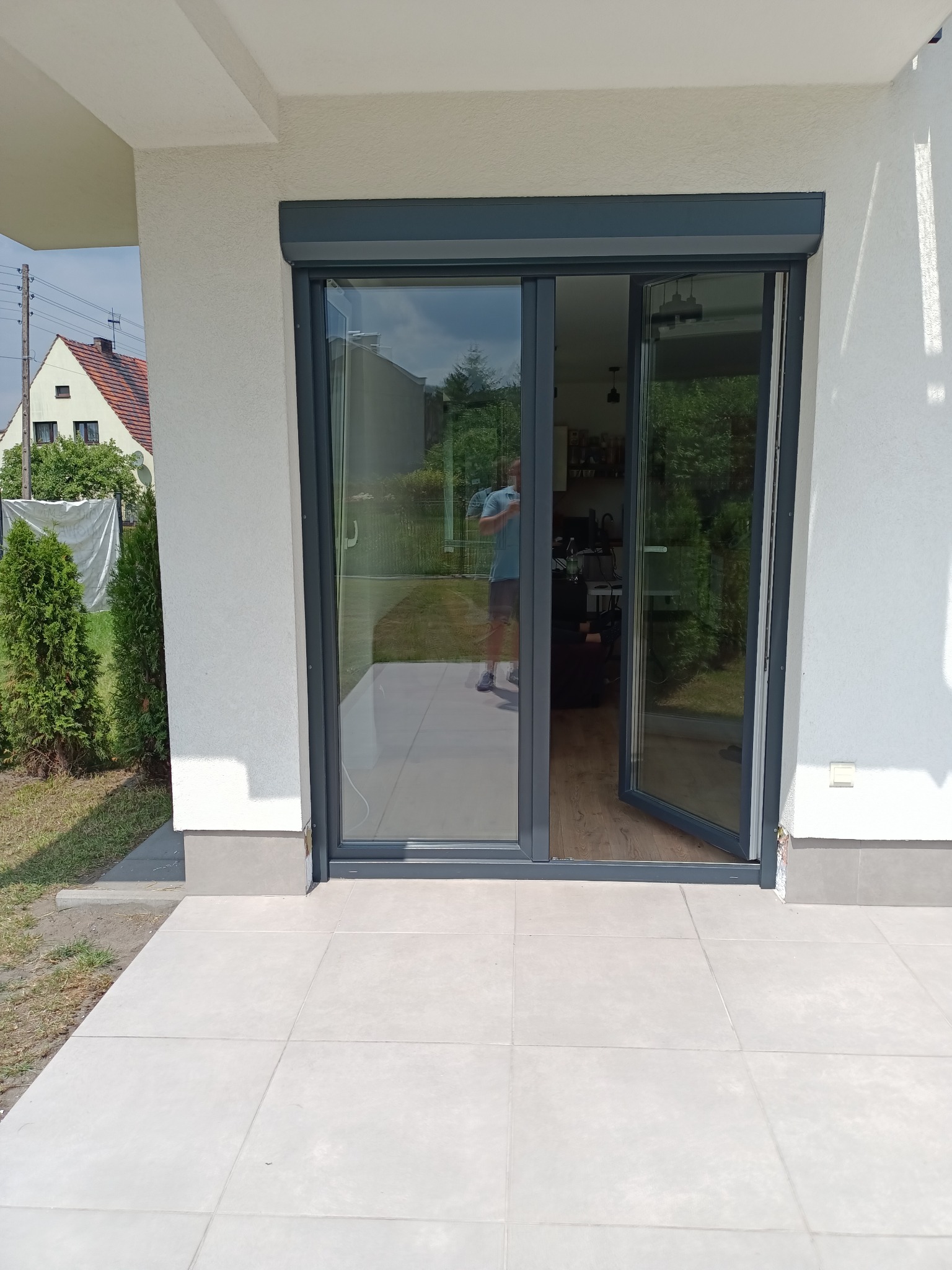 Szare rolety zewnętrzne zamontowane nad przesuwnymi drzwiami tarasowymi, widok z zewnątrz na jasny taras i fragment ogrodu.