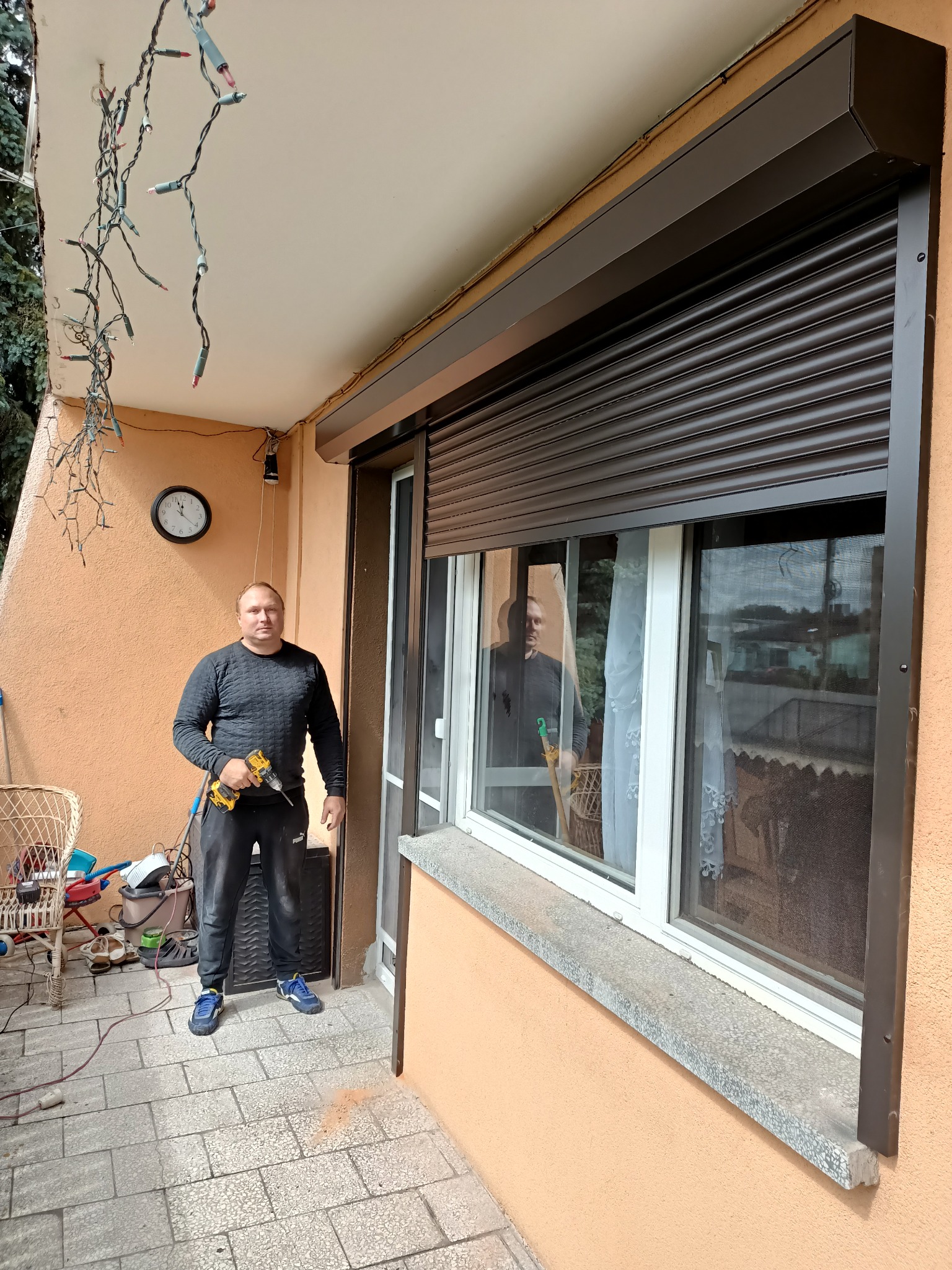 Pracownik z wiertarką montuje brązową roletę zewnętrzną na balkonie z dekoracyjnymi lampkami.