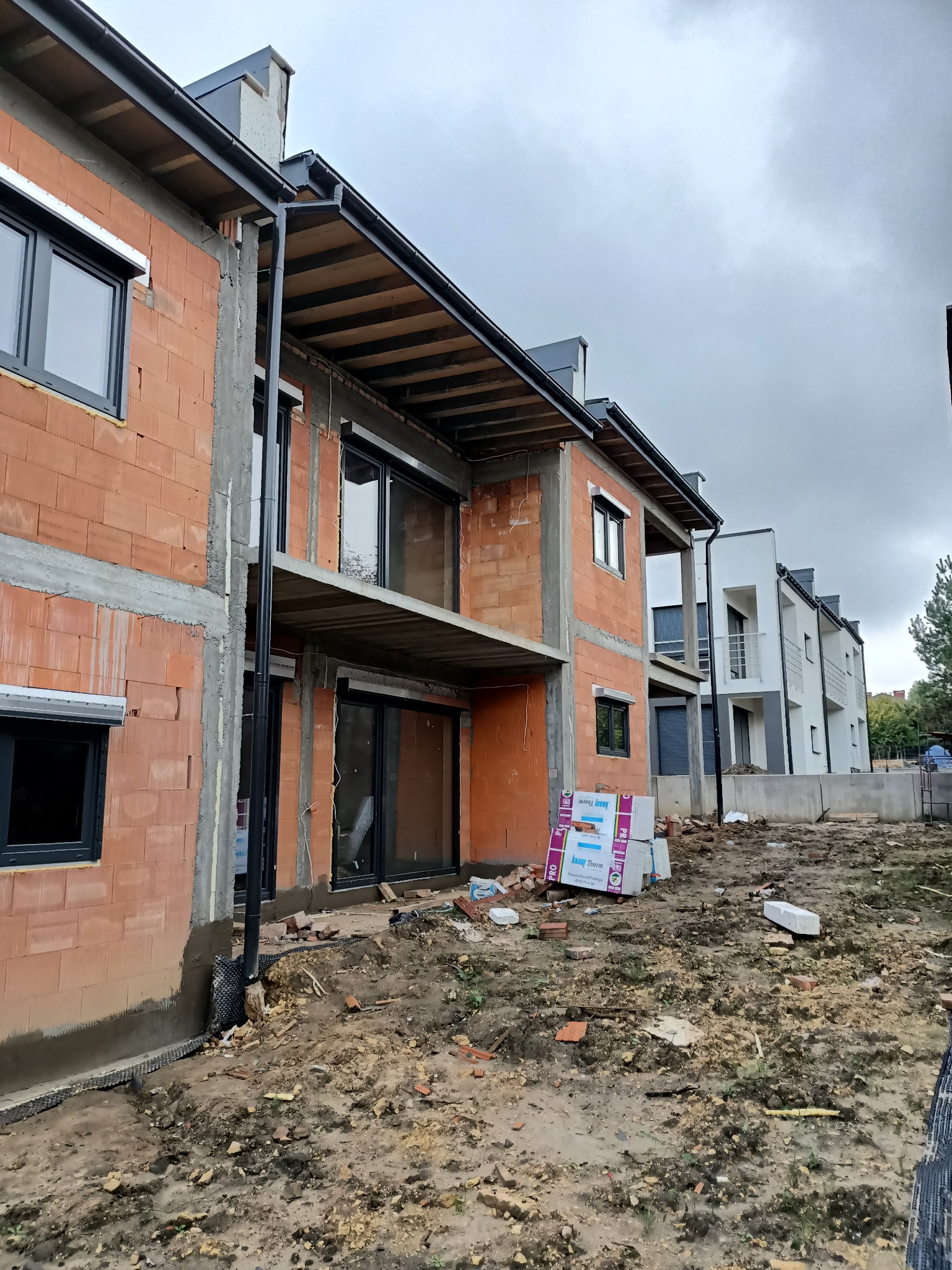 Nowe budynki w stanie surowym otwartym z zamontowanymi oknami i częściowo zamontowanymi roletami zewnętrznymi, widoczne materiały budowlane na placu budowy.