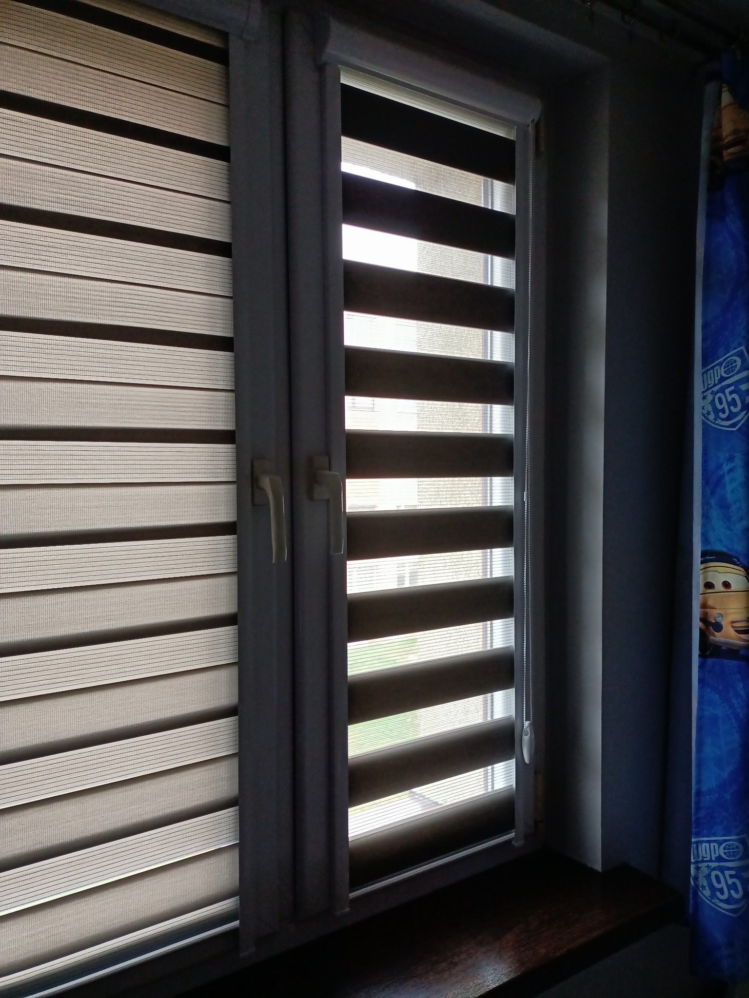 Dwa okna z zamontowanymi roletami typu dzień i noc w kolorach szarości, jedna z łańcuszkiem sterującym, widok z wnętrza pokoju dziecięcego.