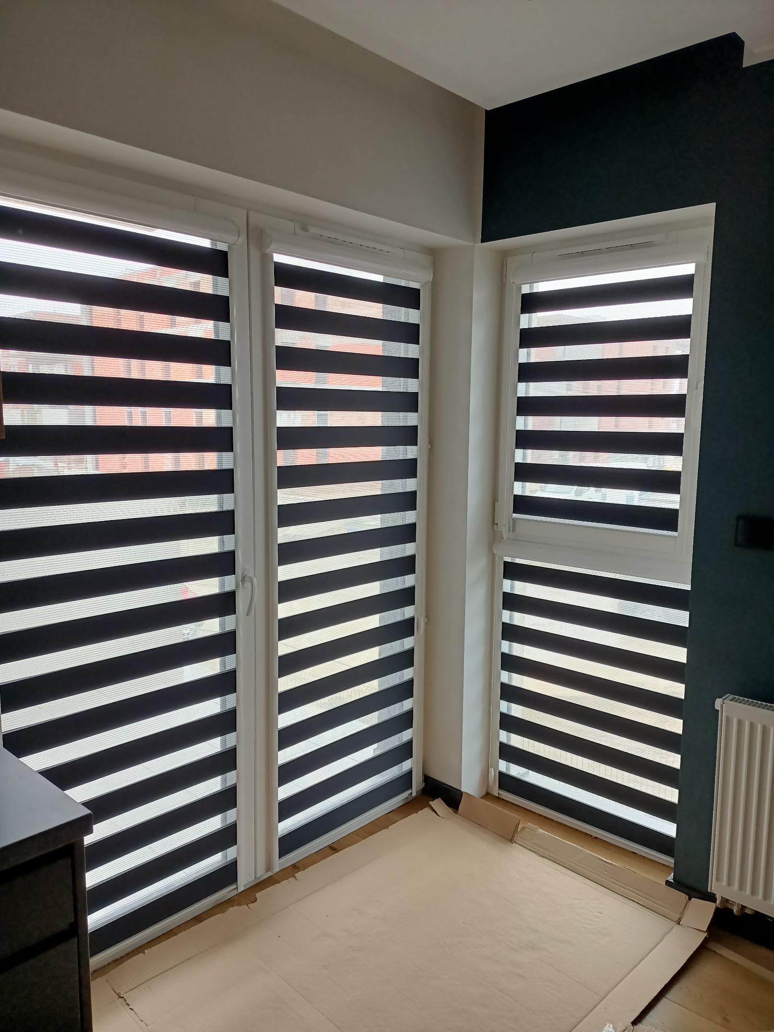 Trzy okna z zamontowanymi roletami typu dzień i noc, widok z wnętrza mieszkania, podłoga zabezpieczona kartonem podczas montażu.