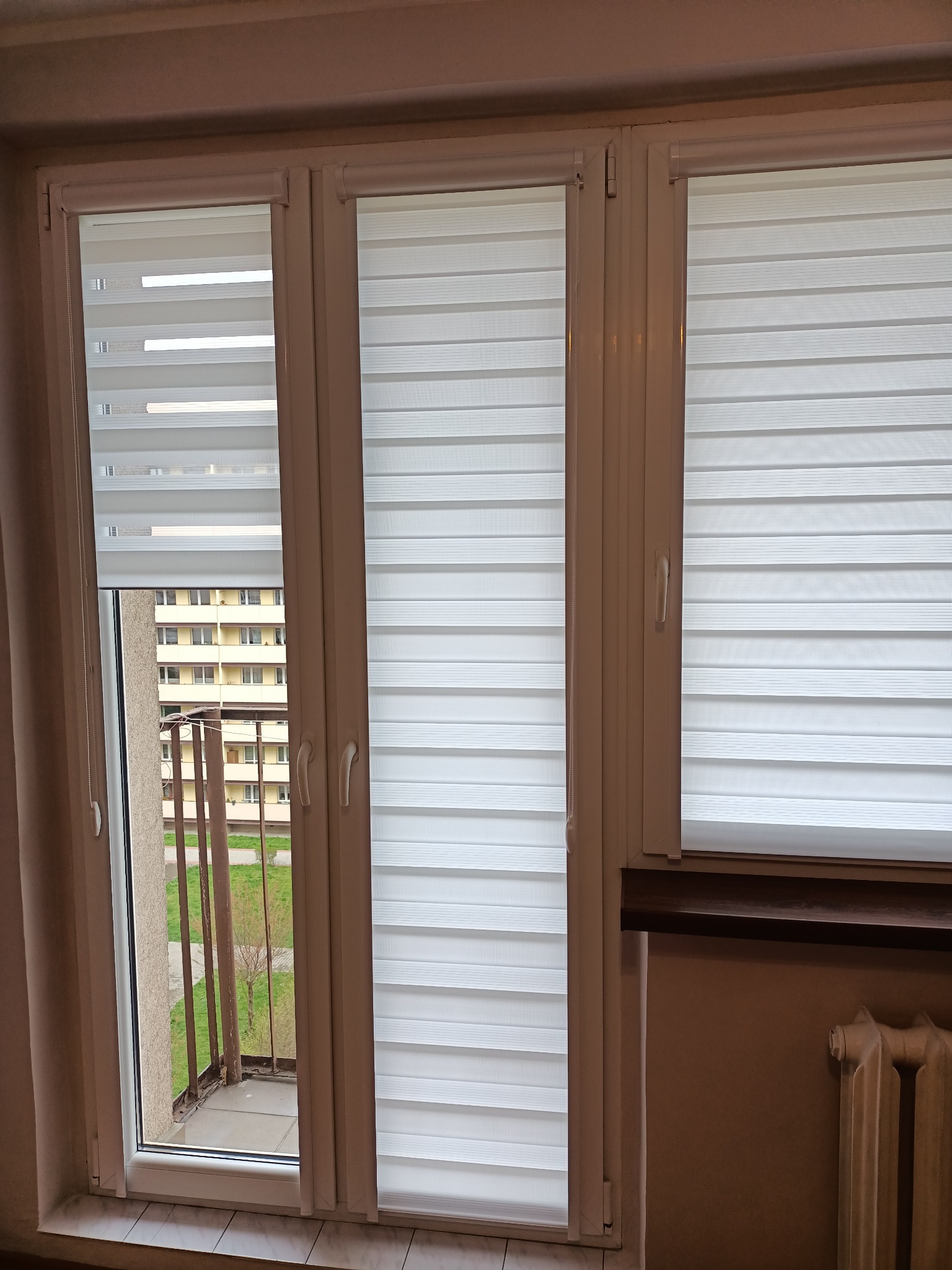 Trzy okna z białymi roletami dzień i noc, widok na balkon i budynek naprzeciwko.