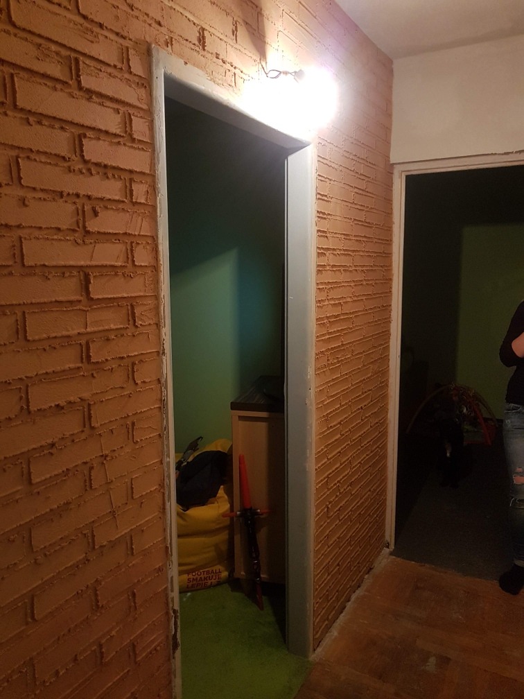 Ściana w korytarzu z fakturą cegły, świeżo po nałożeniu tynku strukturalnego, widoczne drzwi do pomieszczenia z zieloną ścianą i rekwizytem miecza świetlnego.