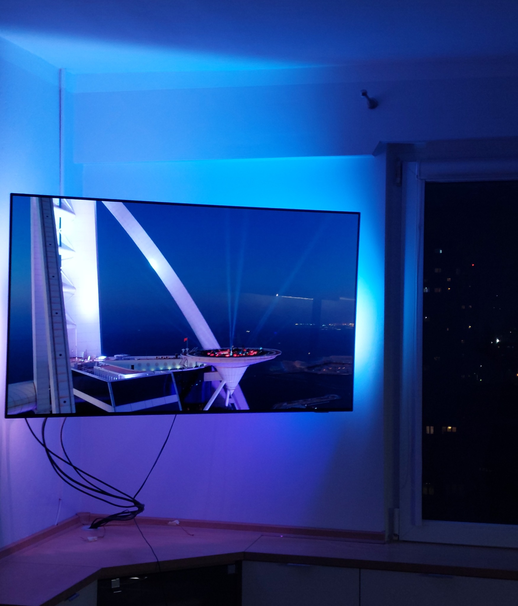 Telewizor zamontowany na ścianie z podświetleniem LED, wyświetlający nocny widok na budynek z platformą widokową, widoczne kable i fragment blatu.