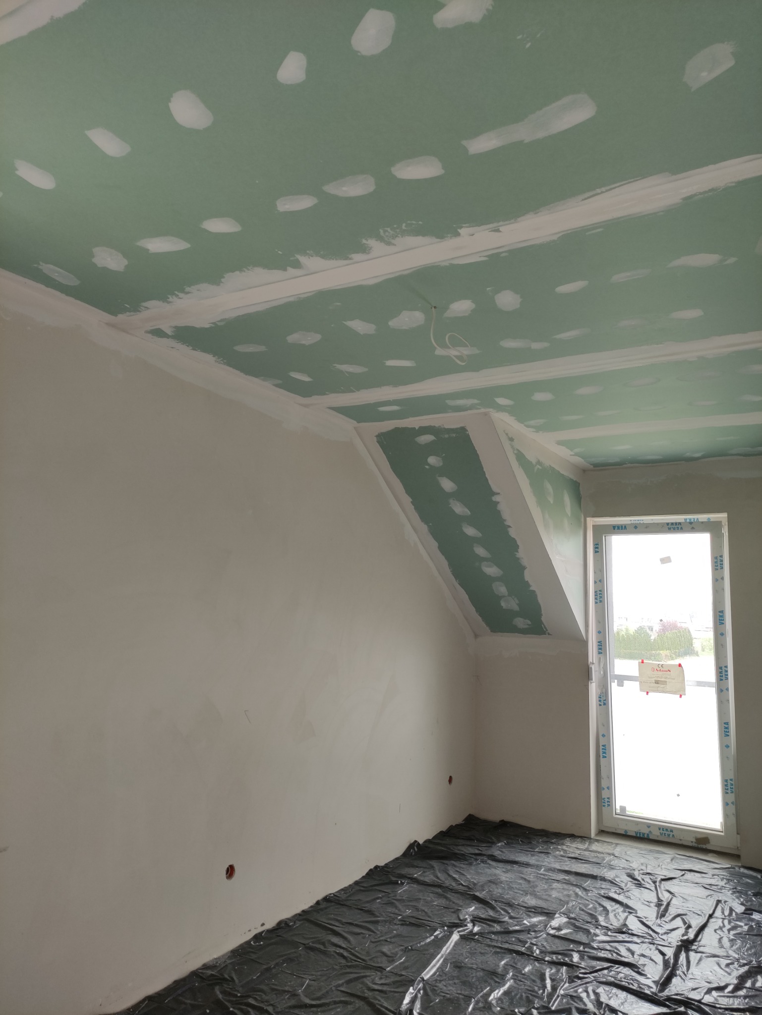 Pokój w trakcie prac wykończeniowych: zielone płyty karton-gips na suficie i skośnej ścianie poddasza, z widocznymi miejscami łączeń i szpachlowania, podłoga zabezpieczona czarną folią.