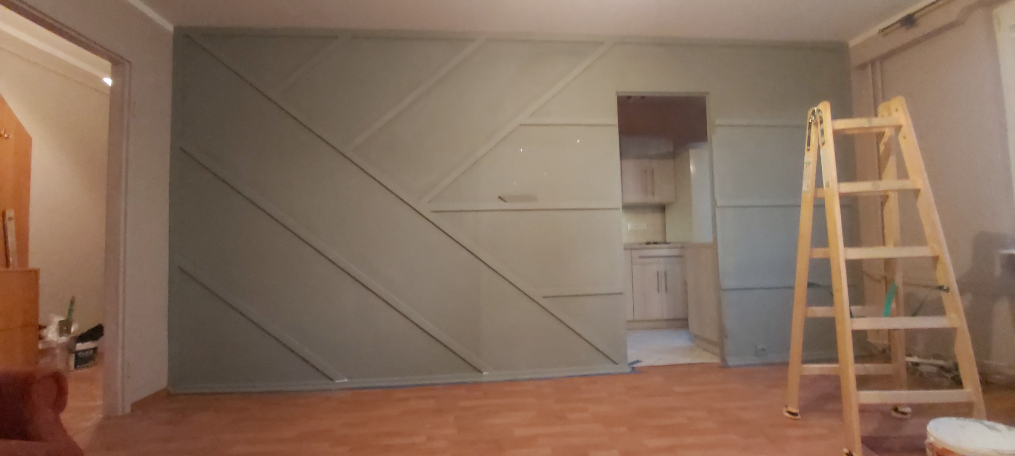 Ściana z dekoracyjnymi panelami w trakcie malowania, widoczna drabina i fragment kuchni w tle.