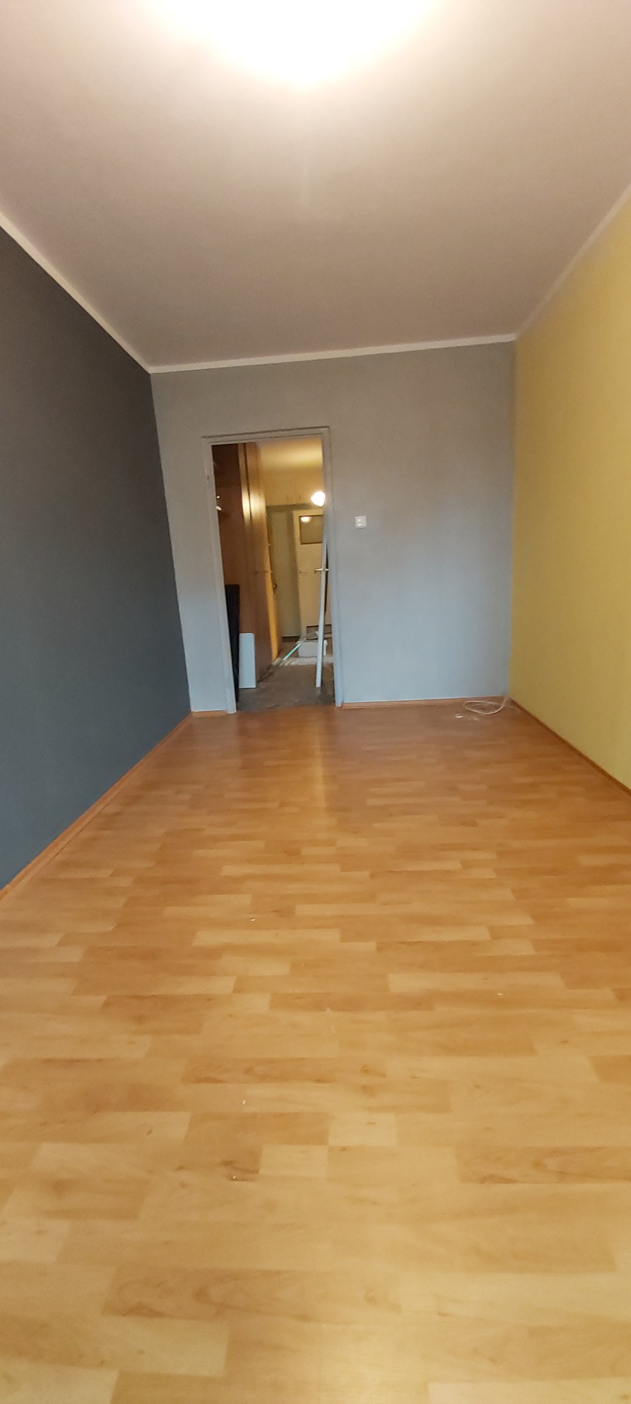 Pokój z nową podłogą laminowaną w odcieniach drewna, świeżo pomalowanymi ścianami w kolorach szarym i żółtym, widok na sąsiednie pomieszczenie przez otwarte drzwi.
