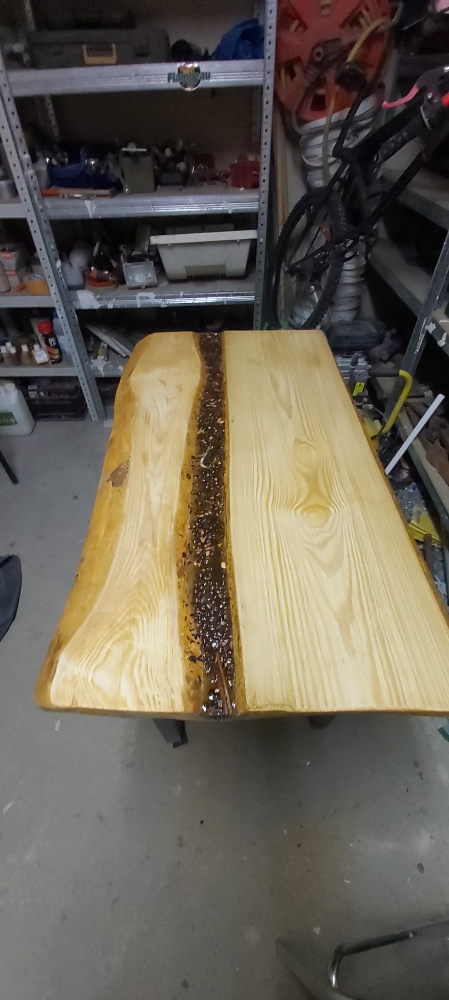 Blat stołu z drewna z żywicą epoksydową, ukazujący naturalne krawędzie i wypełnienie z kamyków w żywicy, na tle warsztatu z regałami i rowerem.