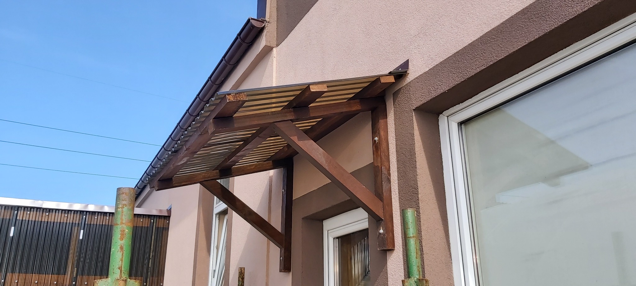 Zadaszenie nad wejściem do domu, wykonane z brązowego drewna i przezroczystego tworzywa, zamontowane na elewacji w kolorze beżowym, widok z boku.