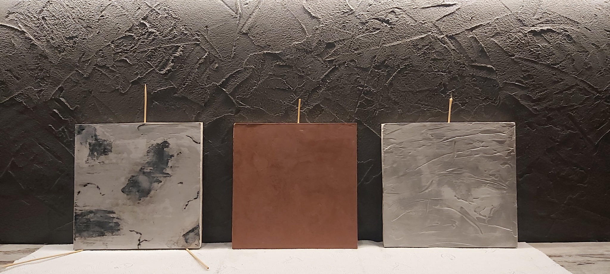 Próbki faktur dekoracyjnych: szara z ciemnymi plamami, brązowa gładka i srebrna z delikatnymi smugami, ustawione na białej powierzchni przed ciemnoszarą ścianą o wyraźnej strukturze.